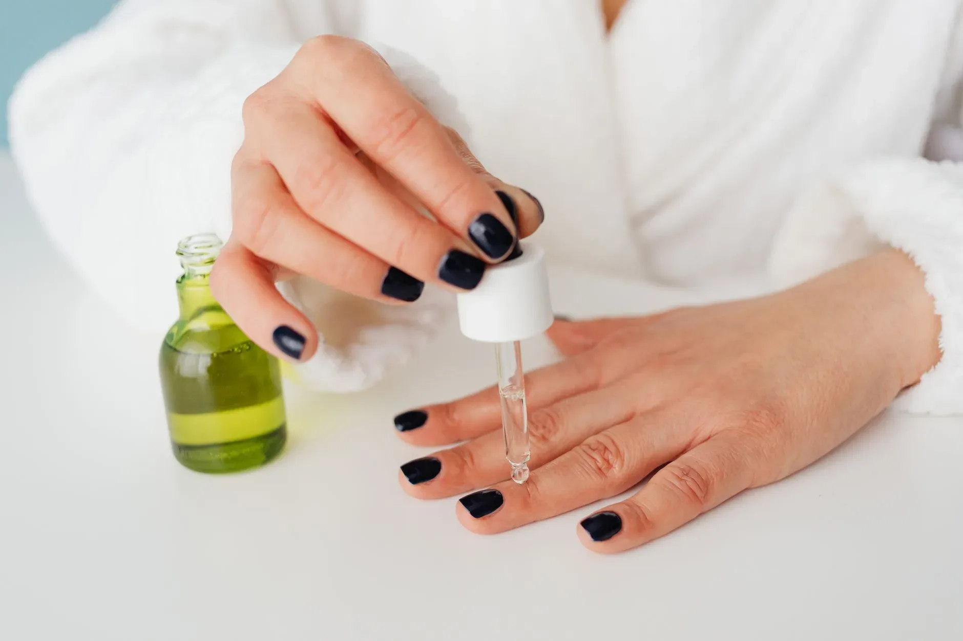Close-up of a woman\'s hands applying serum to nails with a dropper in a bathrobe.